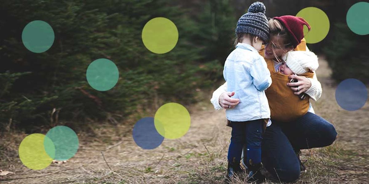 Mother kneels in the woods, holding a baby and comforting her older child as they share a tender moment outdoors.