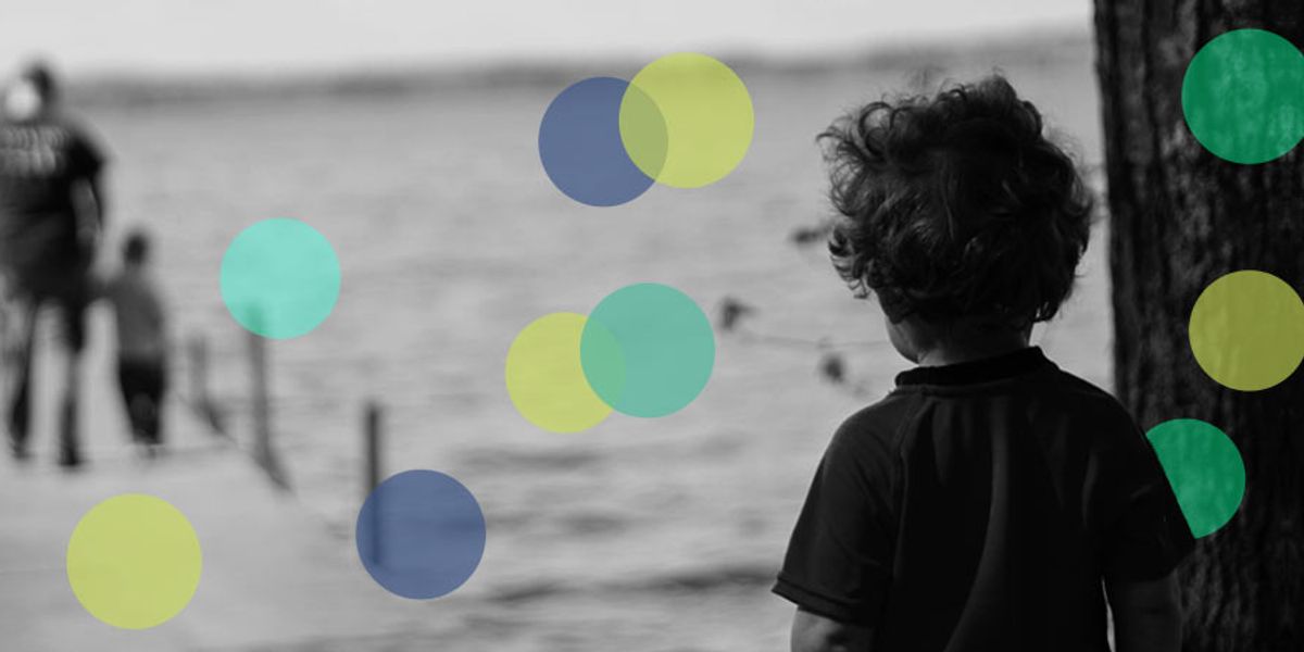 Child standing by a tree looking toward two people walking on a lakeside dock in the distance.