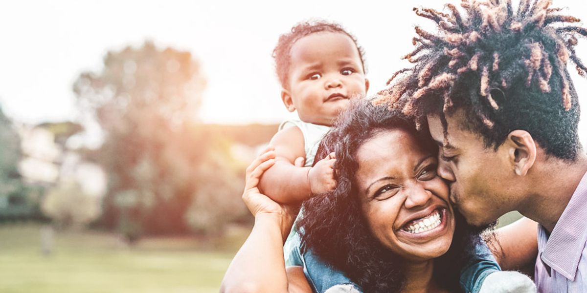 Smiling mother holds a baby while the father kisses her cheek during a joyful moment outdoors.