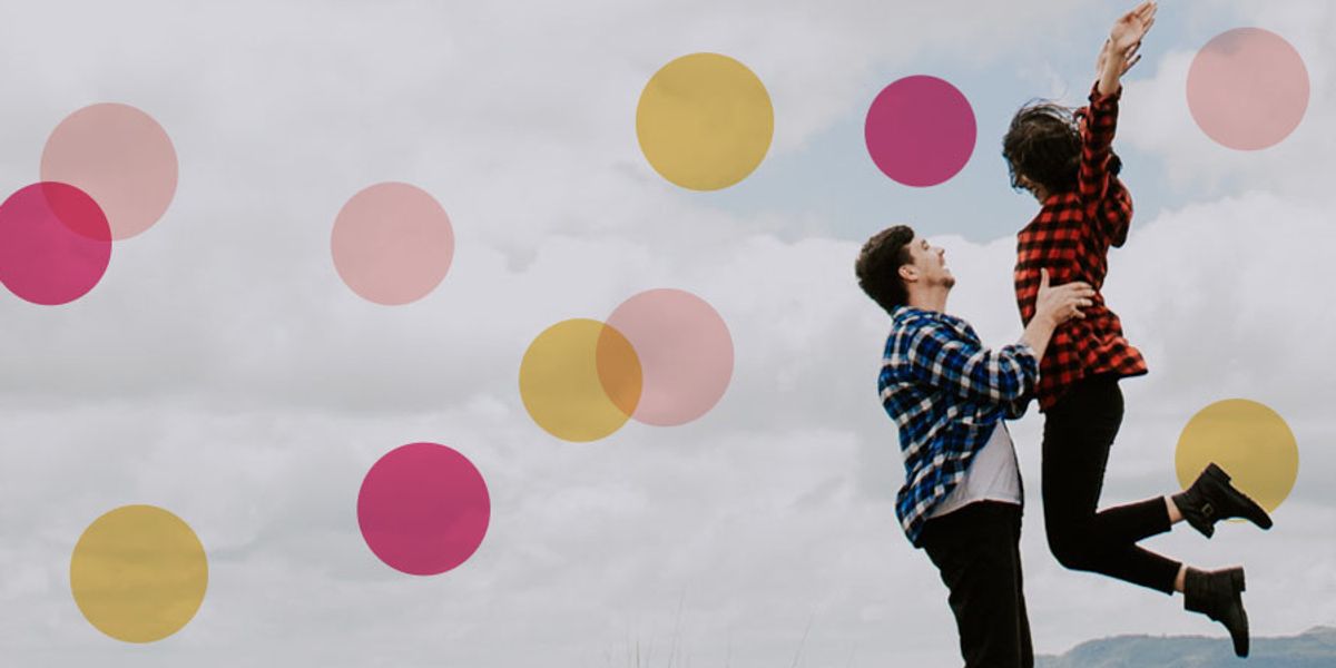 A man lifts his laughing partner into the air outdoors, both wearing plaid shirts against a cloudy sky.