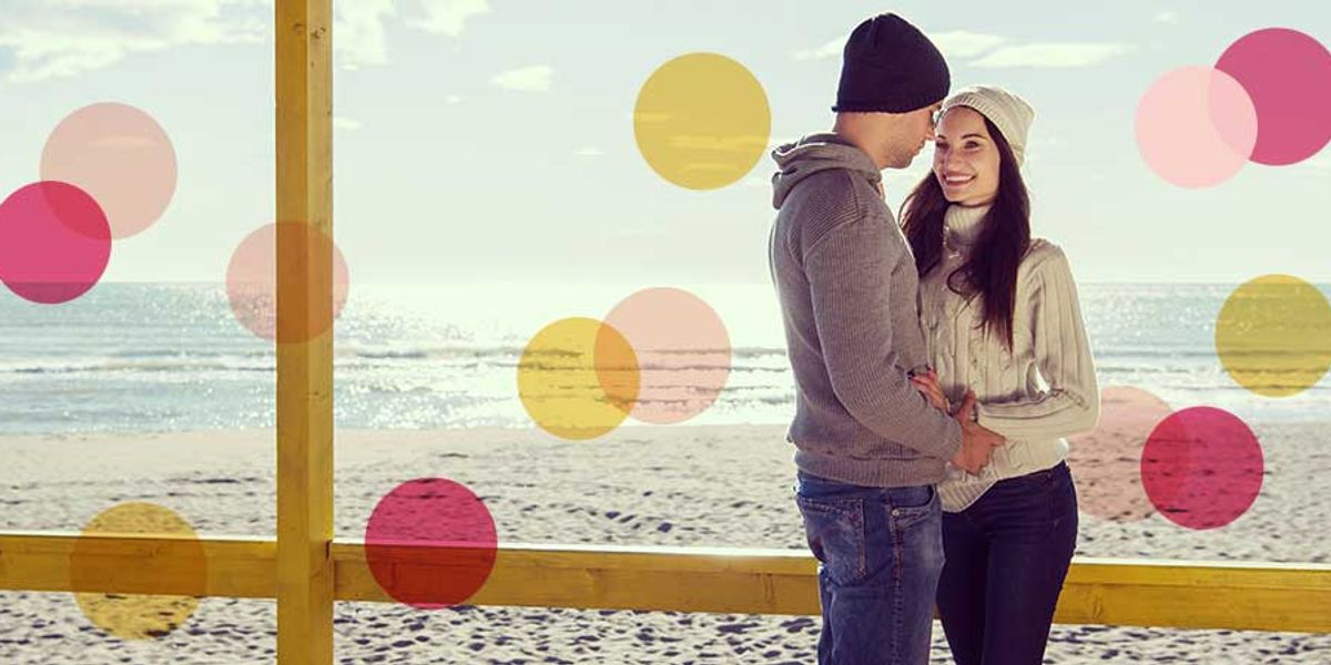 Couple standing together at the beach in winter clothing, smiling and holding each other as soft p