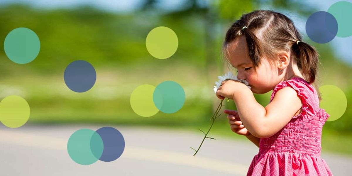 Little girl in a pink dress smelling a white flower while standing outdoors on a sunny day.