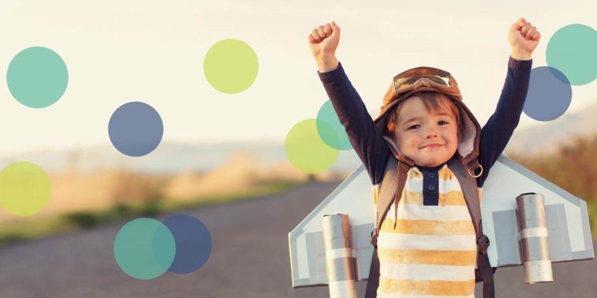 Young child wearing a toy jetpack and goggles raises their arms in excitement while standing outdoors.