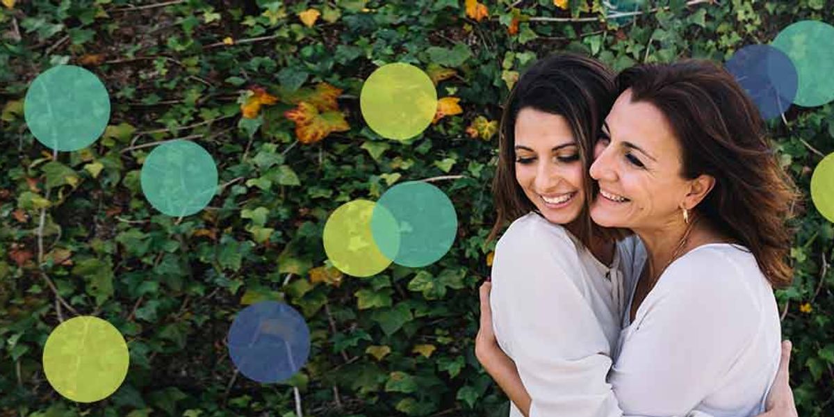 Two women smiling and hugging in front of a leafy green wall outdoors.