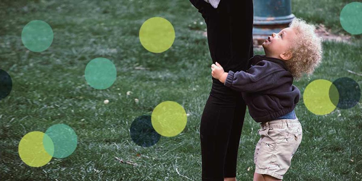Small child hugging an adult’s leg while looking up with a smile in a grassy outdoor setting.