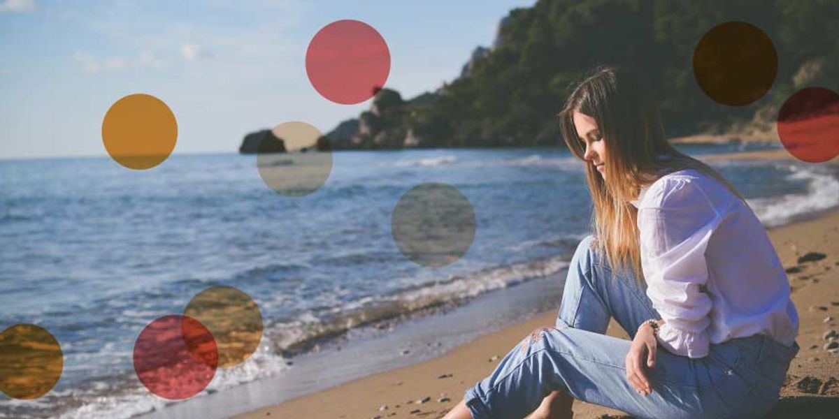 Woman sitting alone on a sandy beach, looking thoughtful as waves reach the shore on a calm day.