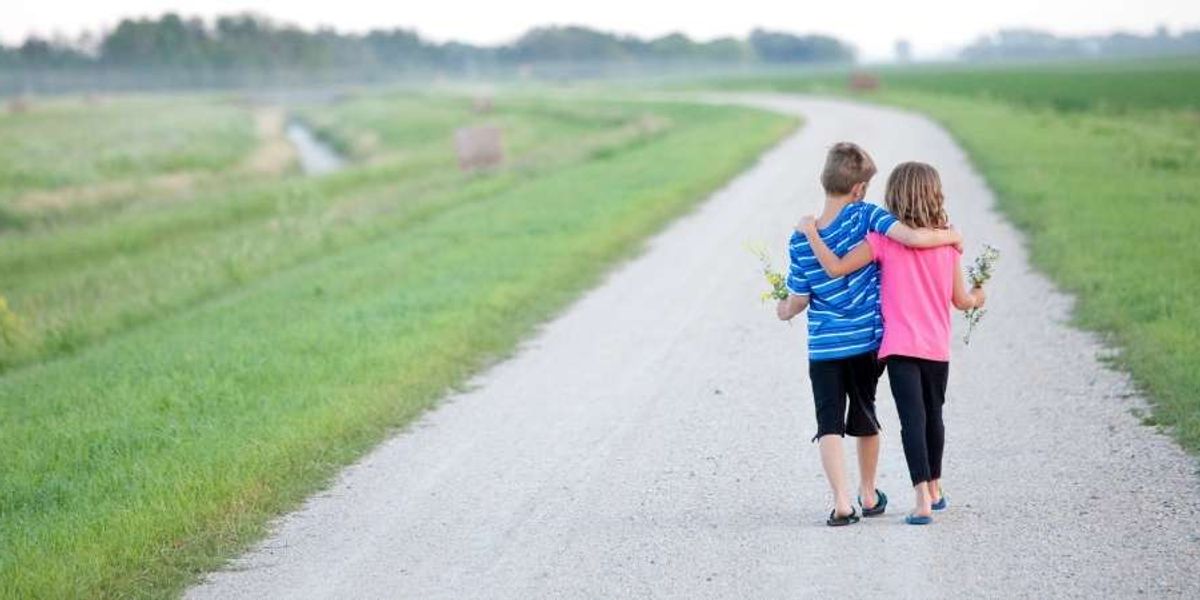 Two children walk down a country road with arms around each other, holding small flowers in their hands.