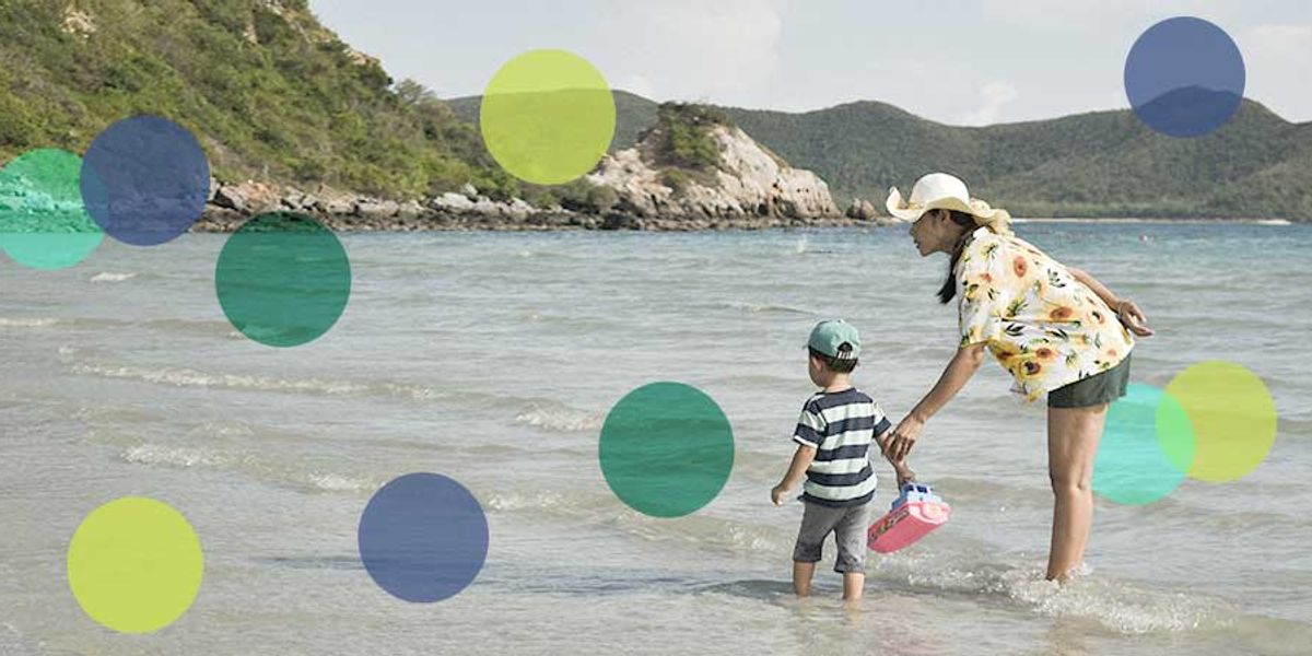 Mother and young son wading in shallow ocean water near a rocky shoreline on a sunny day.