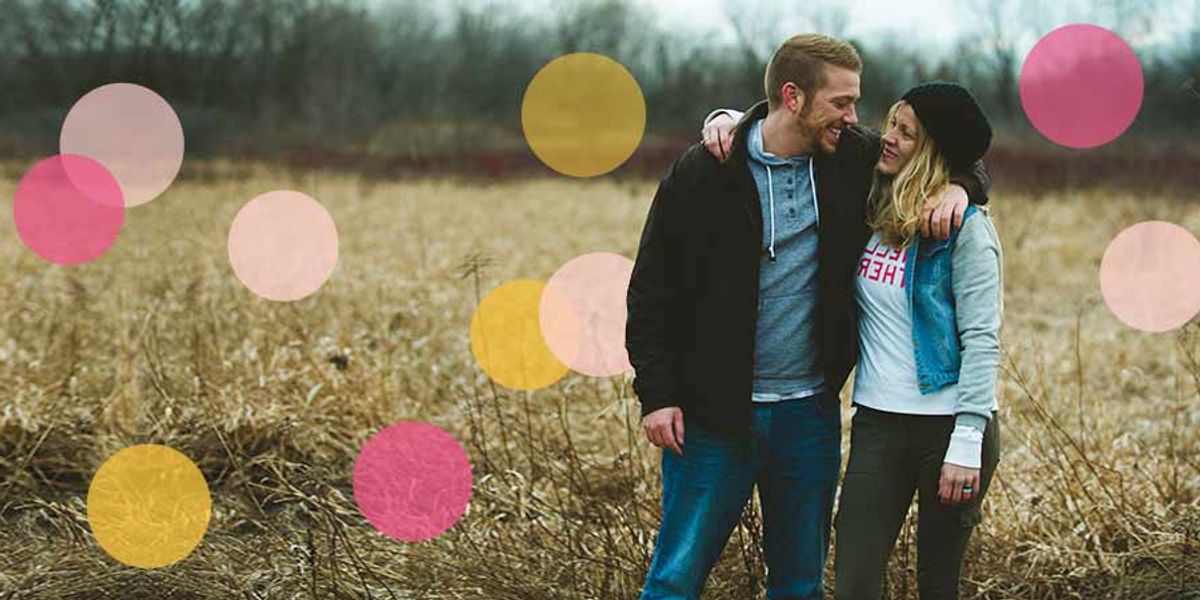 Couple walking through a field on a cloudy day, smiling and holding each other close, with soft pink and yellow circles overlaid on the image.