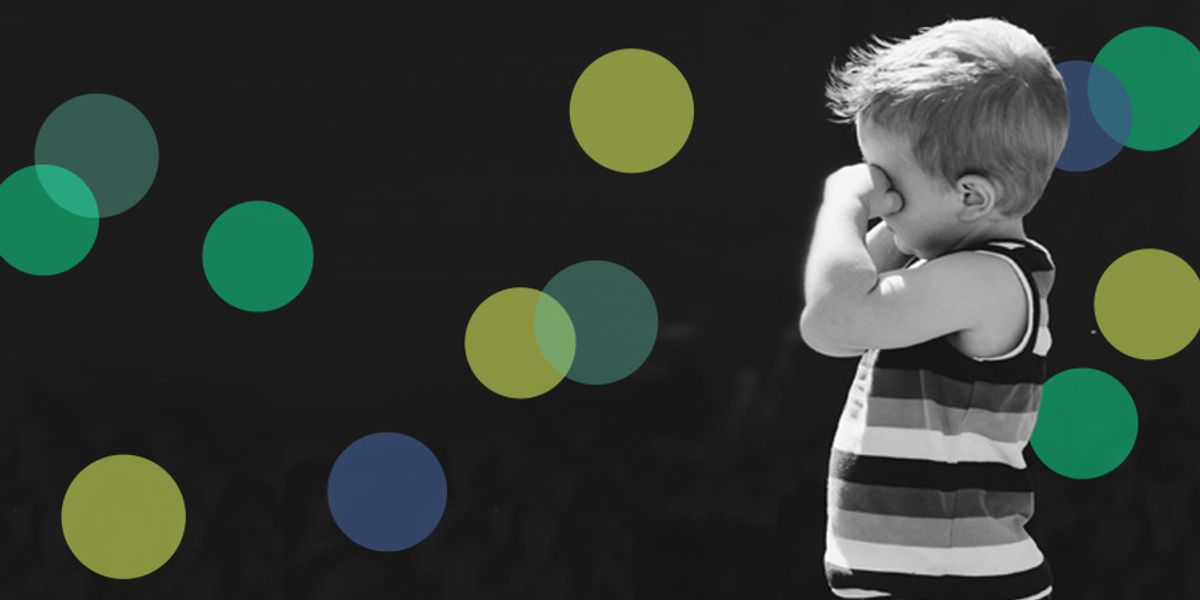 Little boy in a striped tank top rubbing his eyes against a dark background with colorful circles.