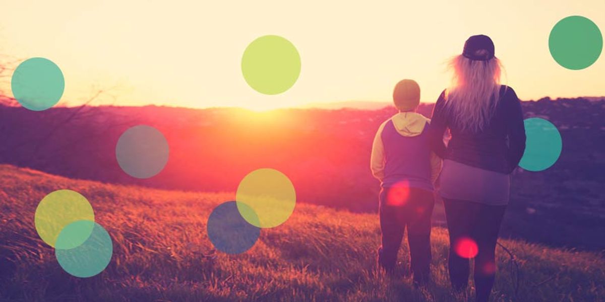 Parent and child standing on a grassy hill at sunset, looking out over the glowing landscape together.