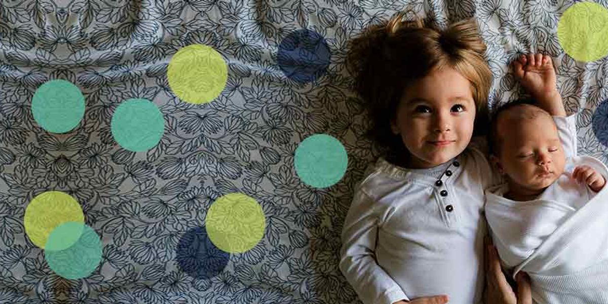 Smiling older sibling lying on a patterned bed beside a swaddled newborn baby, both dressed in white.