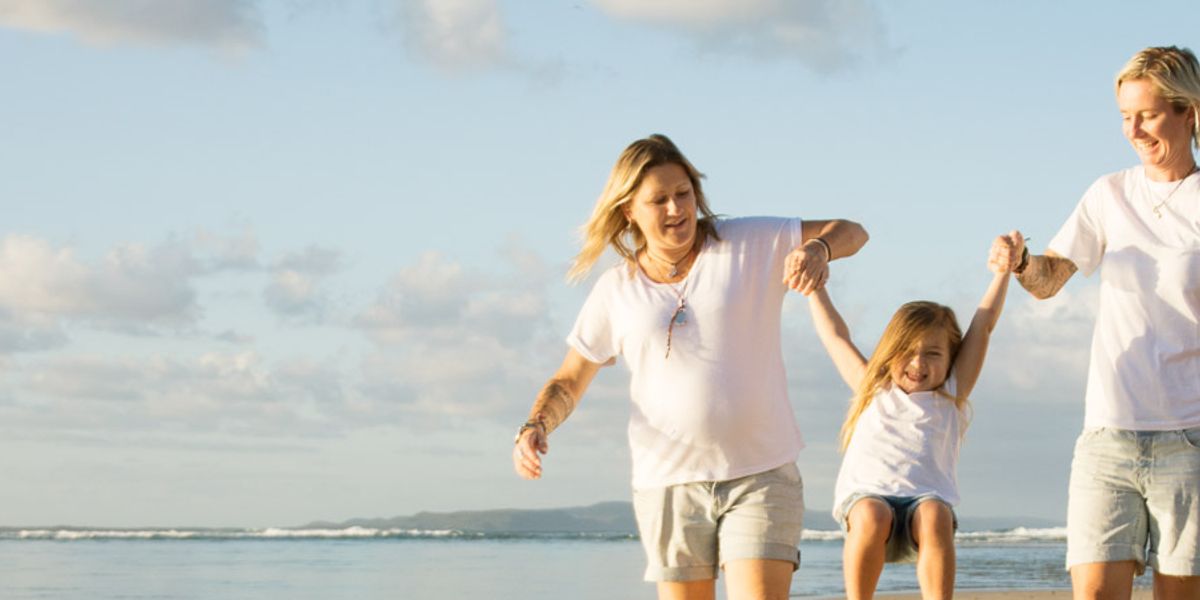 Two women swing a smiling young girl by the arms while walking together along a sunny beach.