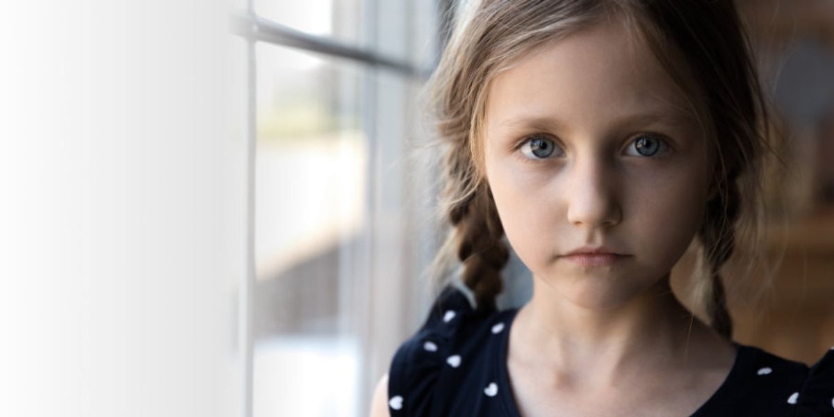 Young girl with braided hair stands by a window, looking serious with soft light on her face.