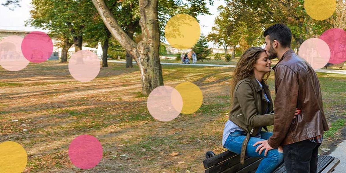 Couple sitting close on a park bench, smiling and leaning in toward each other, with soft pink and yellow confetti circles over the scene.