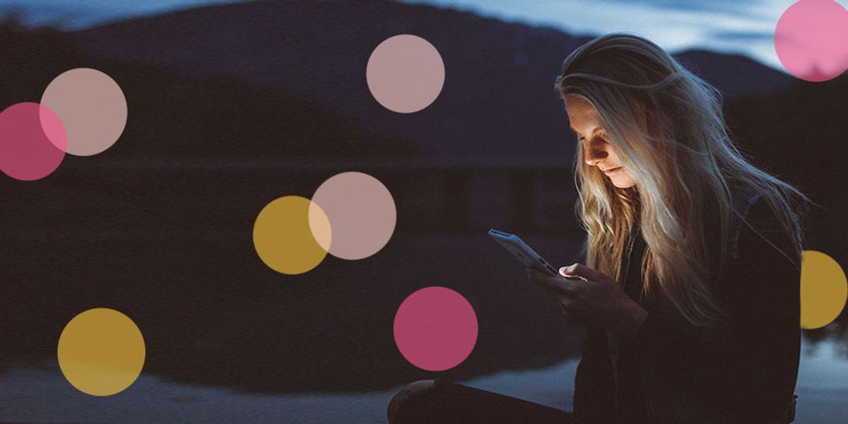 A woman sits by a lake at dusk, illuminated by her phone screen as she looks down and reads.