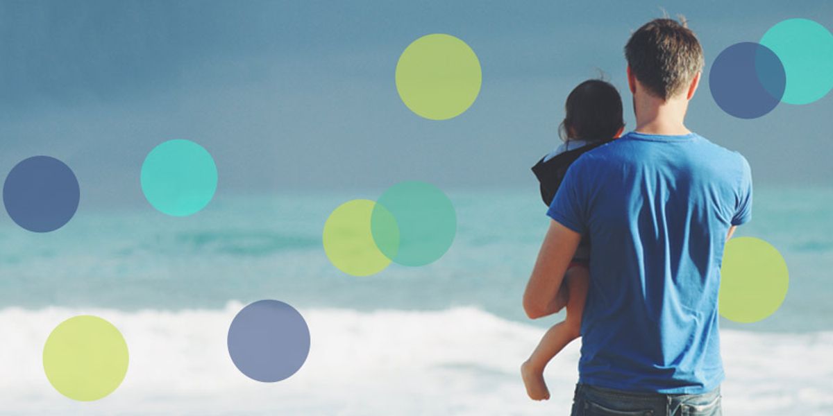 A father in a blue shirt stands at the shoreline holding his child while looking out at the calm, misty ocean.