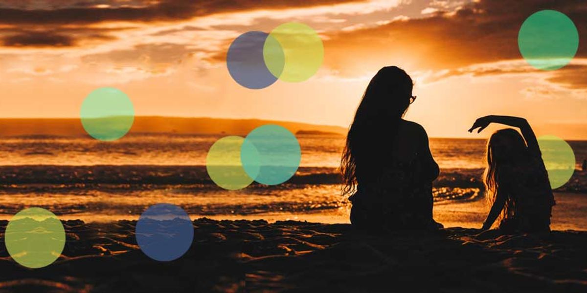 A mother and daughter sit together on the beach at sunset, silhouetted against the glowing sky and waves.