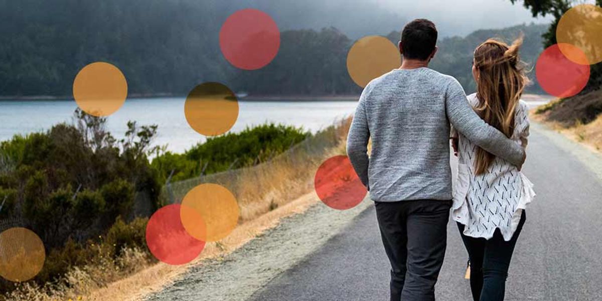 Couple walking arm in arm down a lakeside road with trees and hills in the background