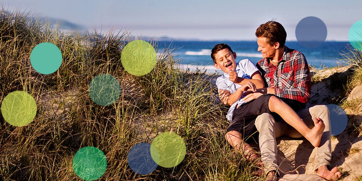 Father and son laughing together on a sandy beach path, sitting among tall grass with colorful circles overlaid.