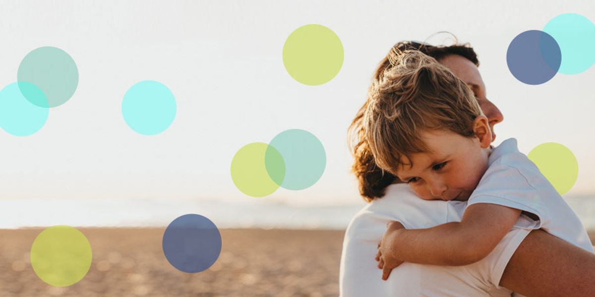Parent holding a tired young boy at the beach at sunset, with soft colorful circles over the image.