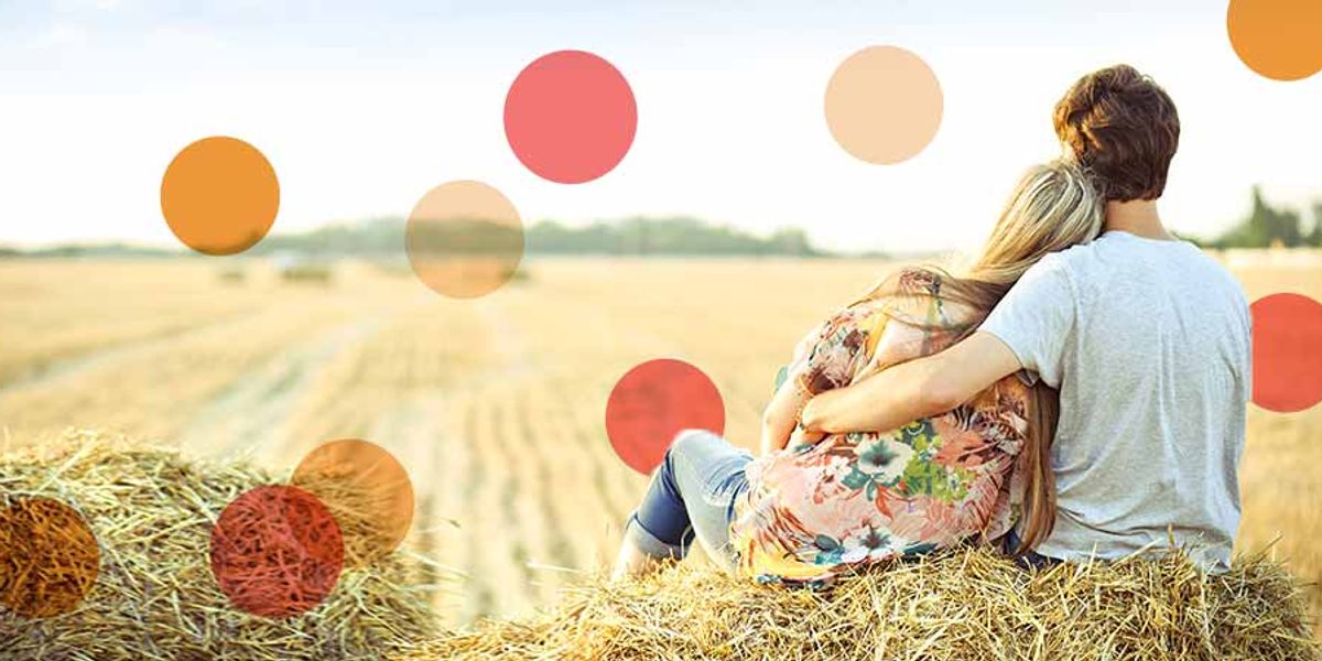 Couple sitting on a hay bale in a sunny field, with the woman leaning on the man’s shoulder