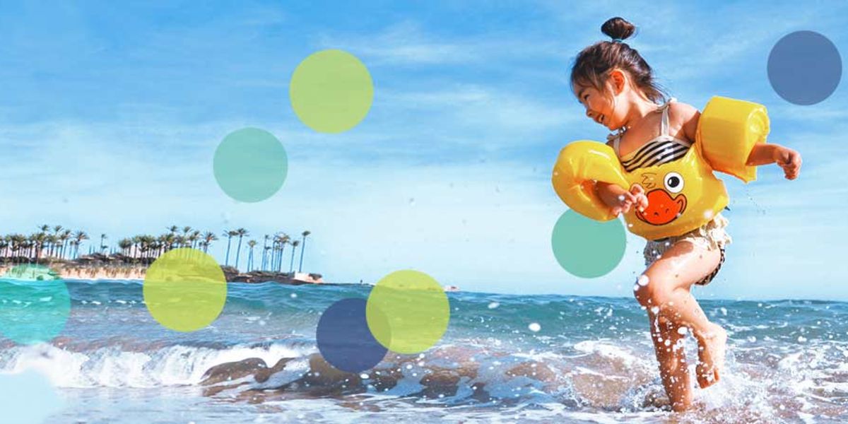 Young girl in yellow floaties splashing in shallow ocean waves on a sunny beach.