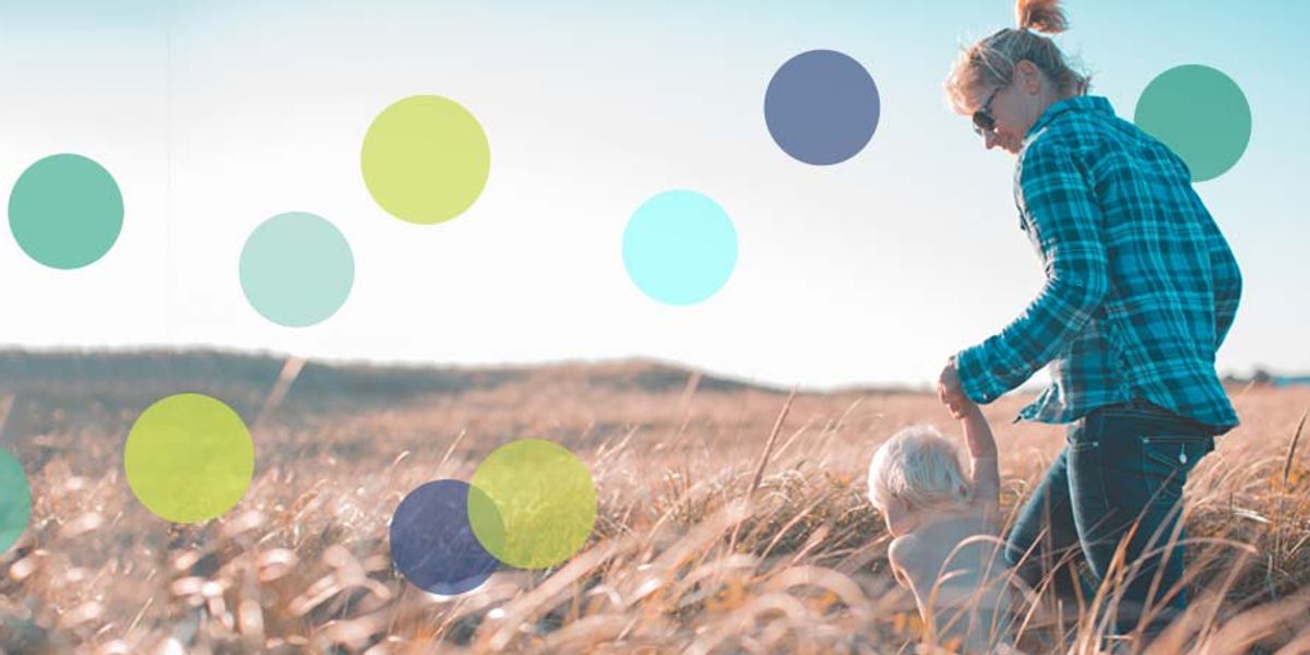 Mother holding her young child’s hand while walking through tall grass on a sunny day.