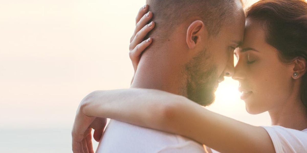 Couple embracing closely at sunset with their foreheads touching, eyes closed, and the sun glowing behind them.