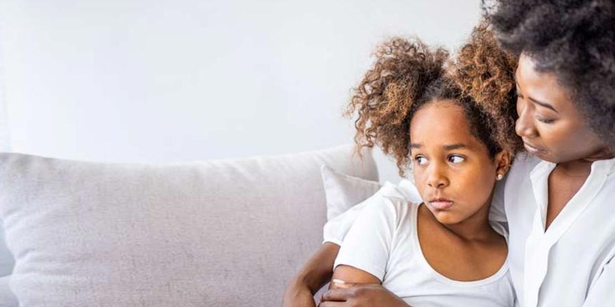 Mother comforting her upset daughter on a couch, holding her close as the child looks away.