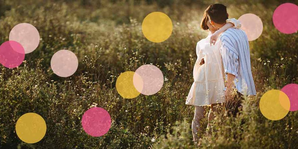Couple embracing in a sunlit field, standing close together among tall grass with soft pink and yellow circles overlaid.