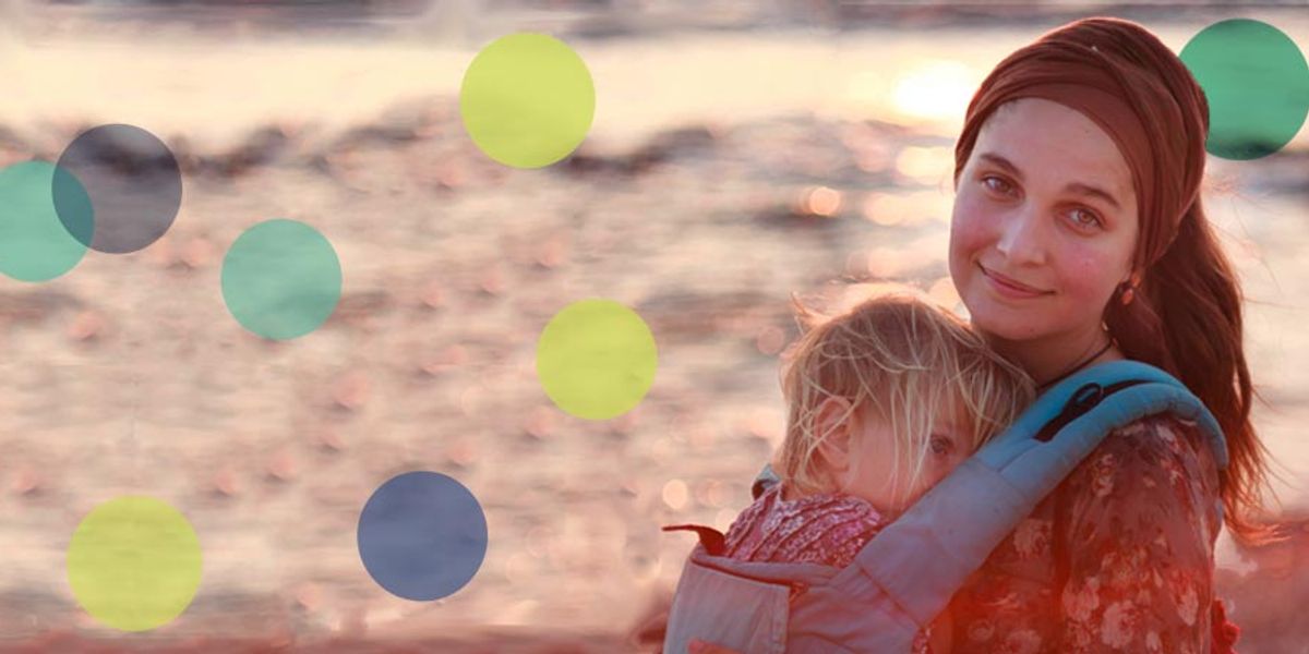 Mother holding her sleepy child in a carrier by the water at sunset, both calm and close.