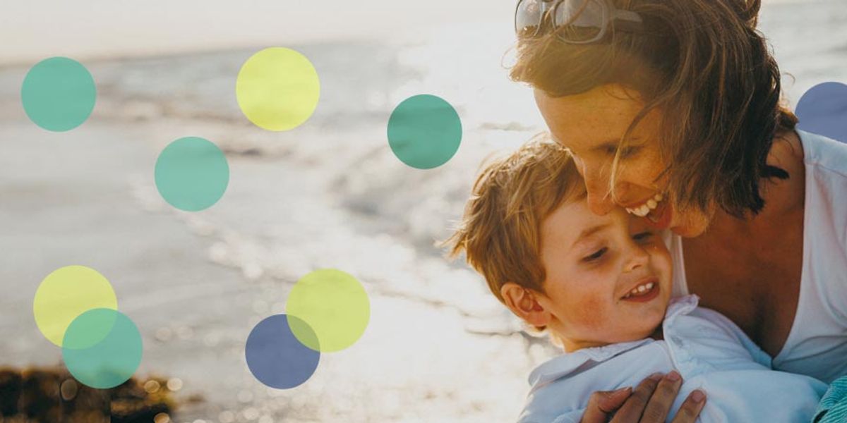 Mother laughing as she hugs her young son on a sunny beach, ocean waves in the background.