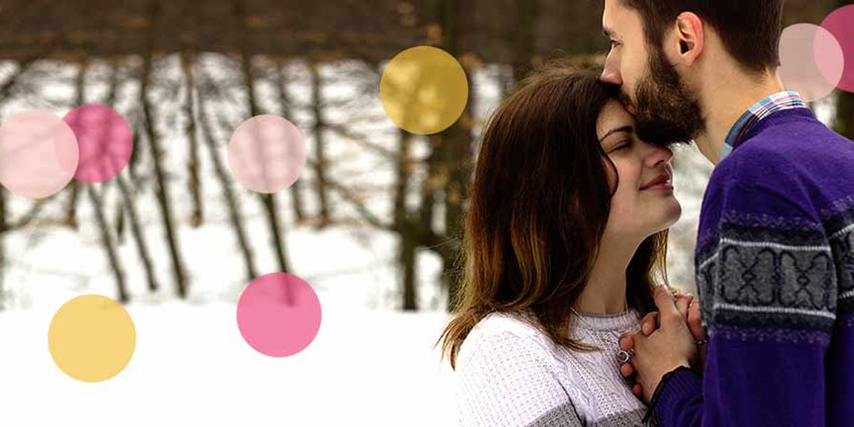 Couple standing in the snow as he kisses her forehead and they hold hands, with soft colorful circles overlaying the scene.
