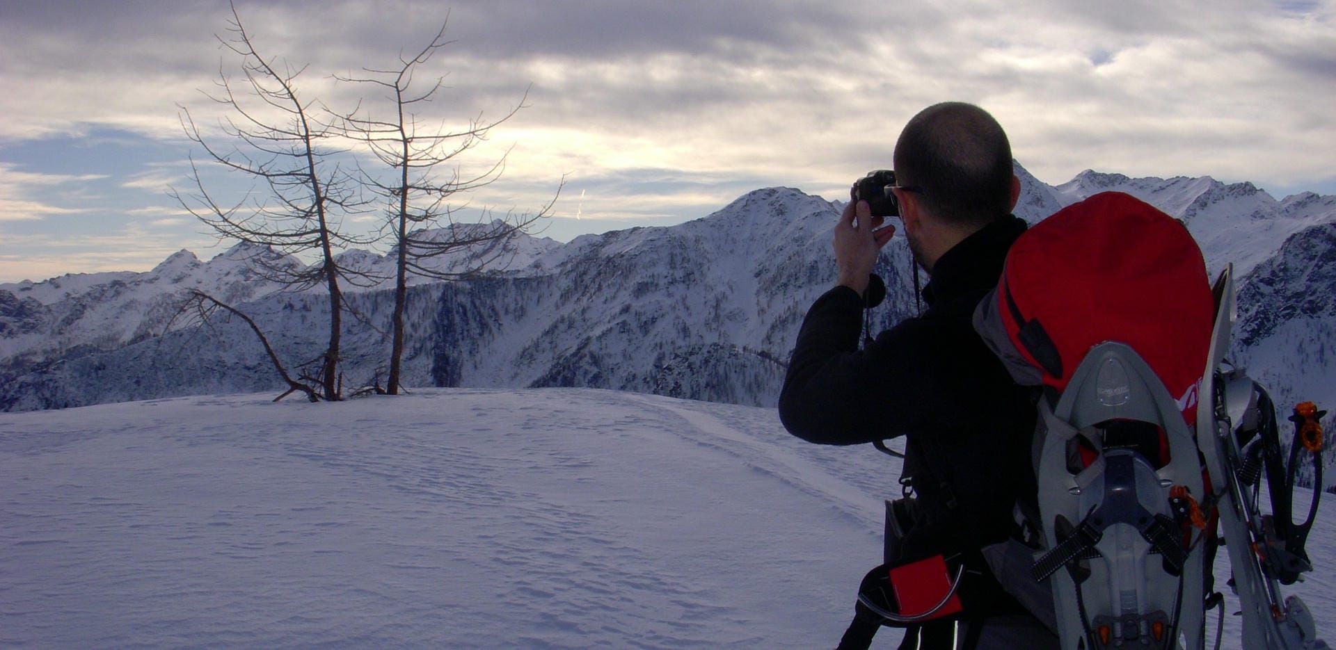 A man in snowy mountains wearing outdoor walking gear and taking a picture of the scene with a camera.