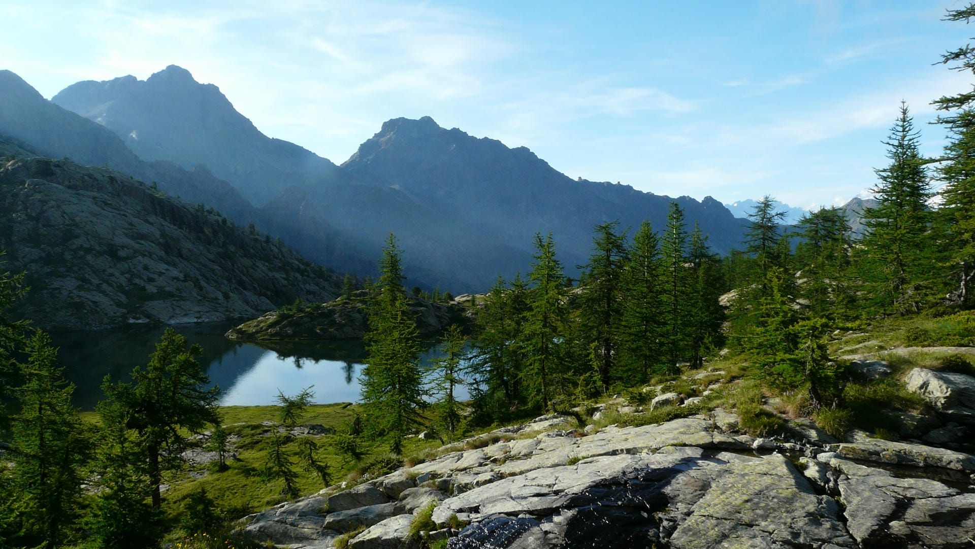 A natural landscape photo showing mountains, pine trees and a lake.