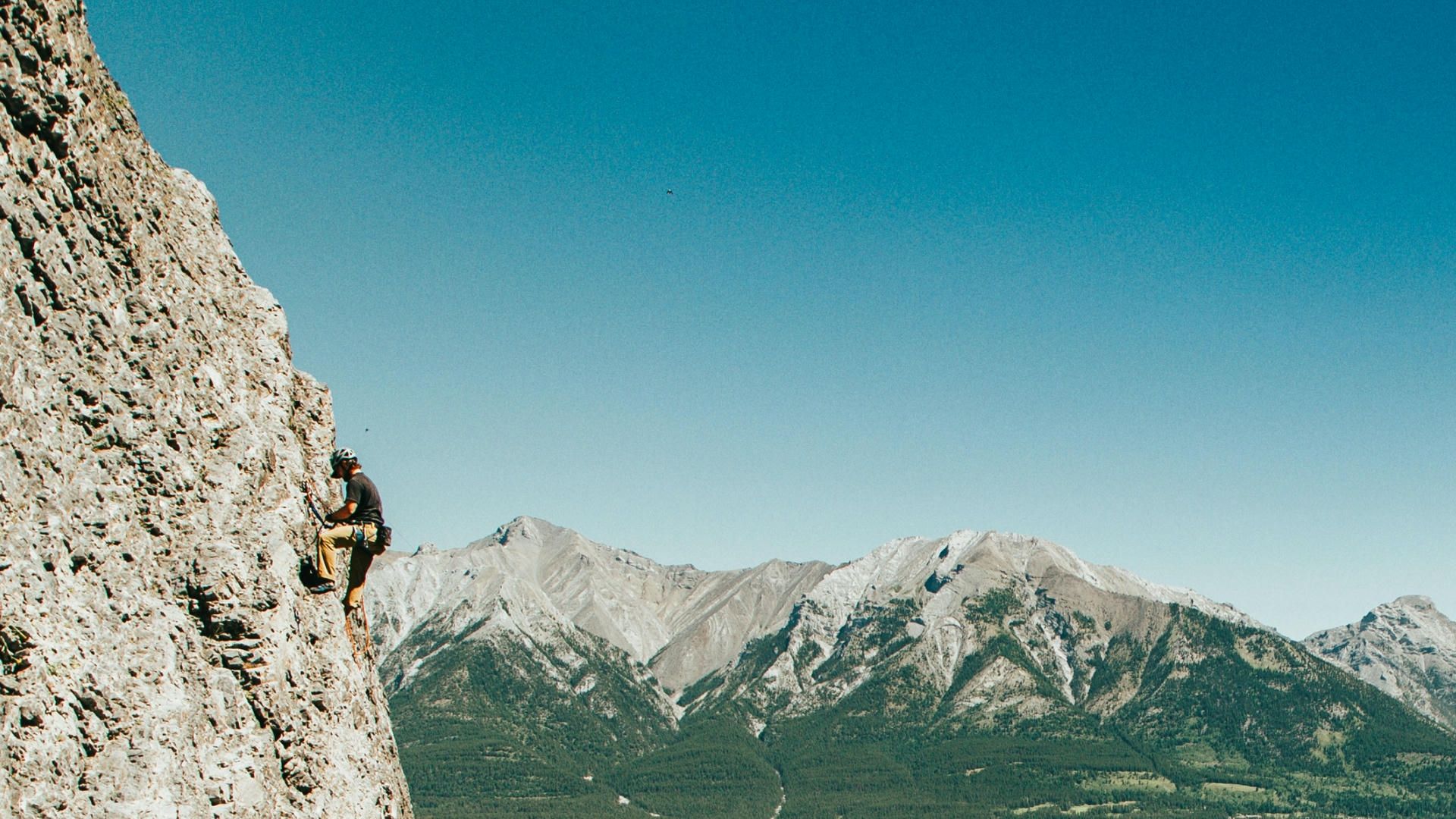 An outdoor photo of a man climbing a mountain with rope and other mountains in the background.