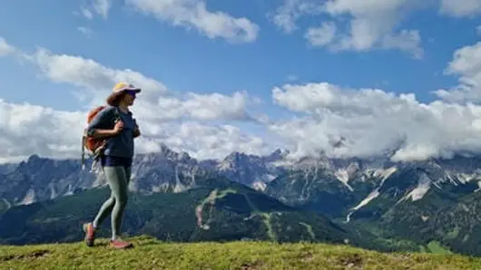 Rocky peaks shrouded in clouds with lush woodland hugging their flanks stand in the background, a woman walks across a grassy alpine path in the foreground.