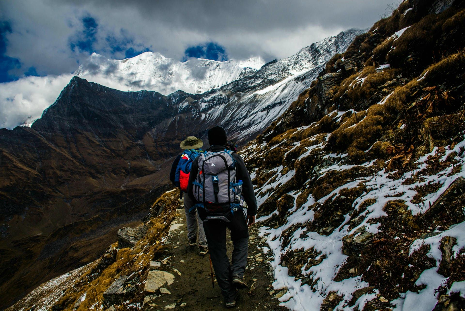 Two mountaineers hiking along a snowy cliff edge wearing outdoor gear like functional backpacks, hats and walking boots.