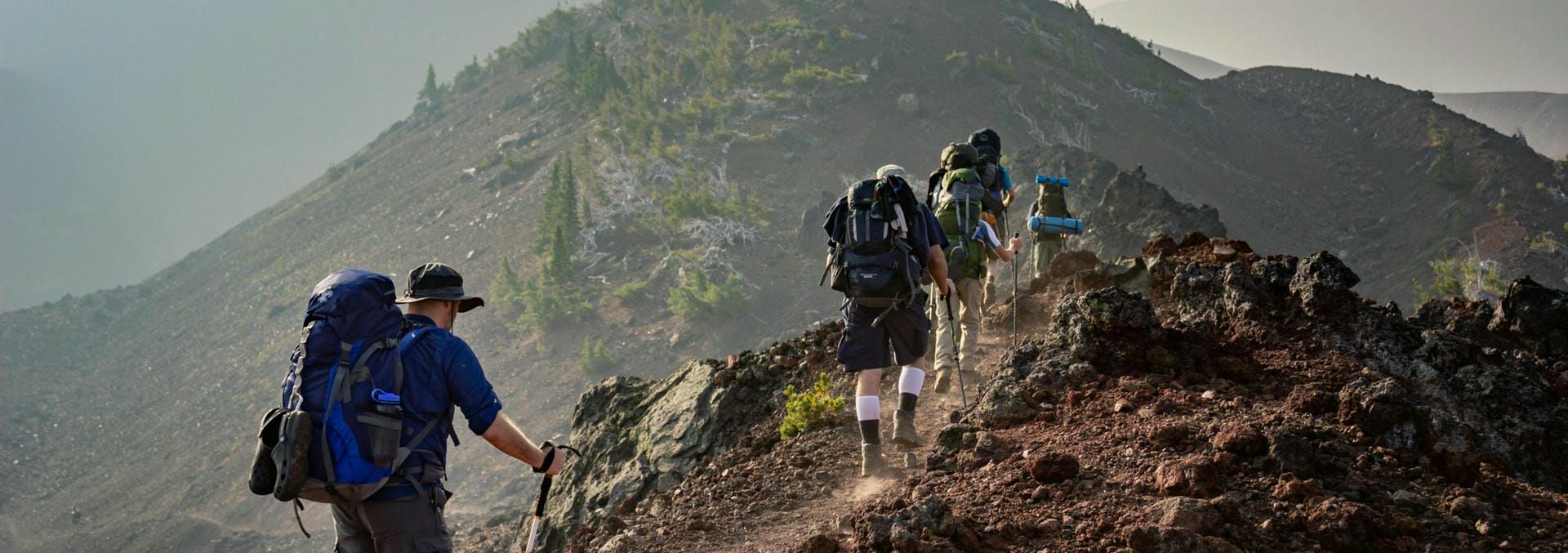 Several hikers wearing outdoor gear such as backpacks, hats and walking poles hiking on the top of a mountain in a line.