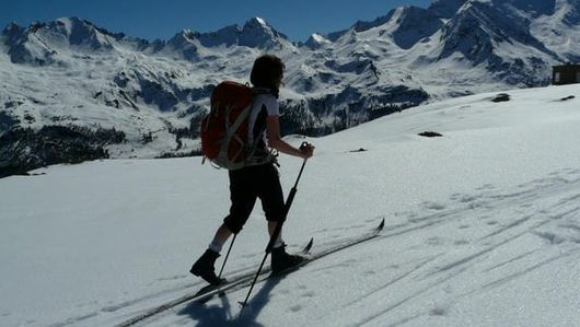 Photo of cross country skier moving across a snowy plateau with a snow-covered mountain range in the background.