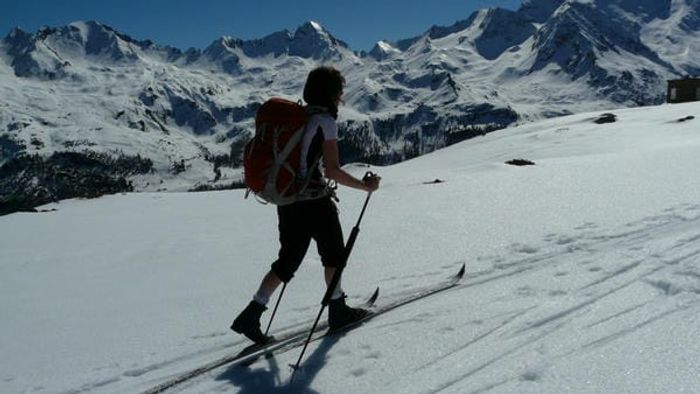 Photo of cross country skier moving across a snowy plateau with a snow-covered mountain range in the background.