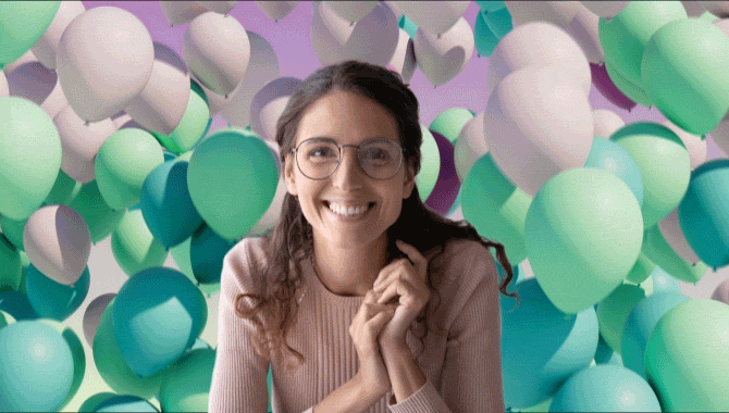 Woman in pink sweater and glasses smiling in front of green, white, pink balloons rising