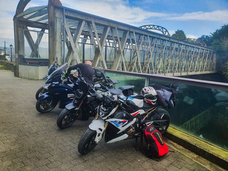 Touring motorcycles parked beside a historic iron lattice bridge during a rest stop on a group ride