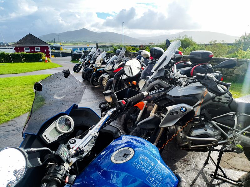 Row of touring motorcycles beaded with rain parked beside a harbour with mountains in the distance
