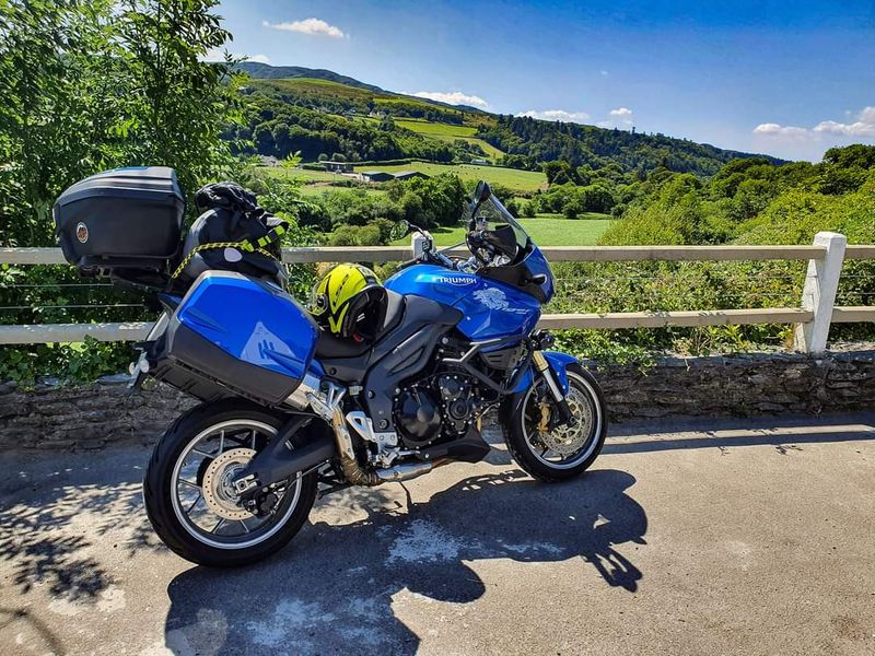 A blue Triumph Tiger touring motorcycle loaded with luggage parked by a country fence overlooking green Irish hills