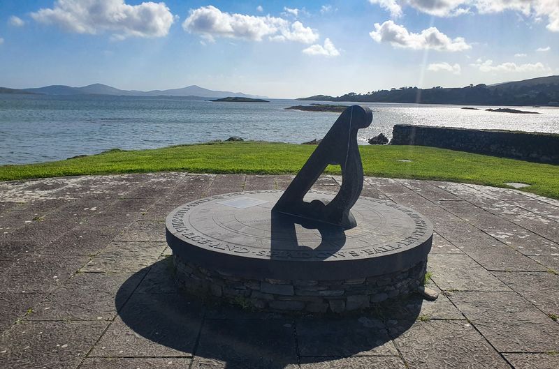 Carved stone sundial monument overlooking a sunlit bay dotted with islands on the southwest Irish coast