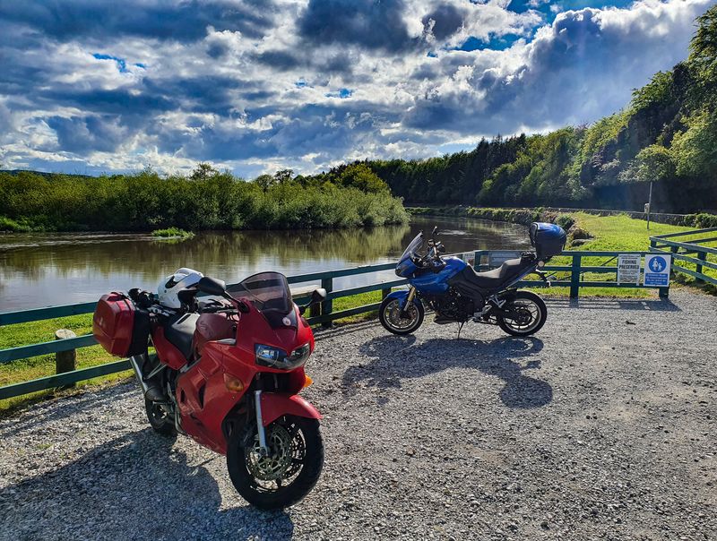 Two touring motorcycles parked at a quiet riverside stop under dramatic clouds in rural Ireland