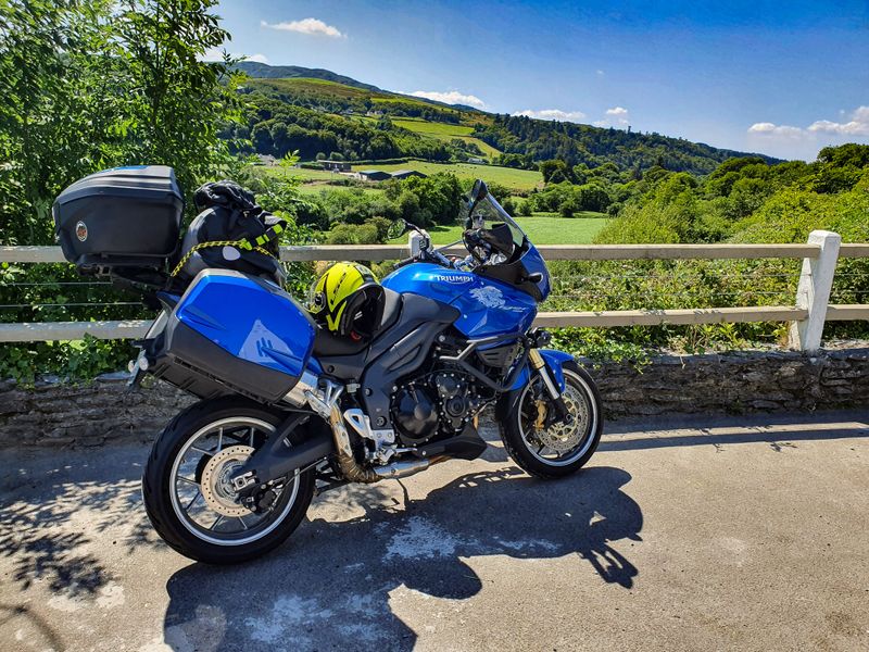 Fully loaded Triumph Tiger touring motorcycle parked on a stone bridge overlooking green Irish hills