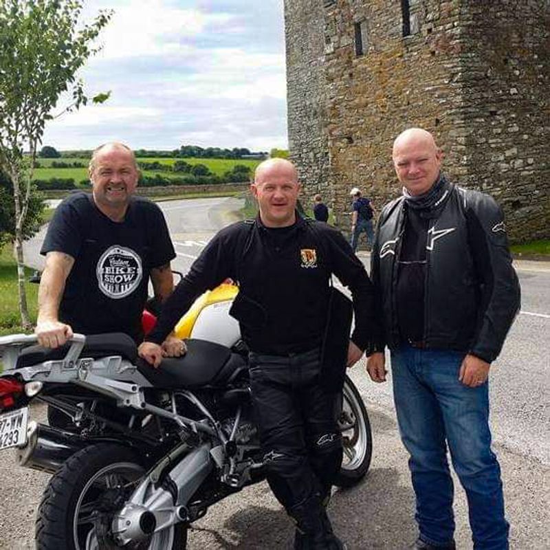 Three motorcyclists smiling beside a BMW GS in front of an old Irish stone castle tower